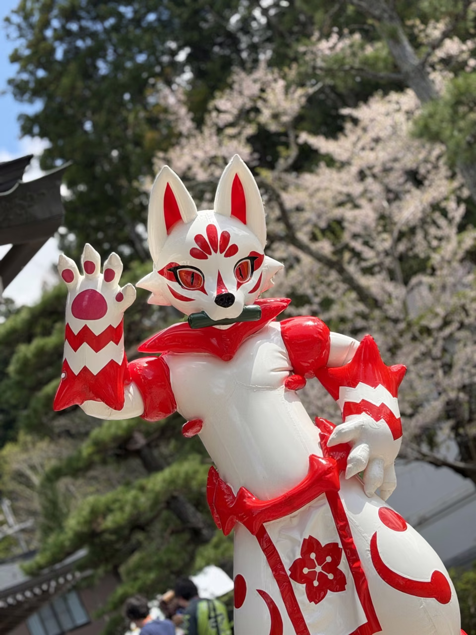 White GUO series latex kitsune with red markings waving in front of a temple