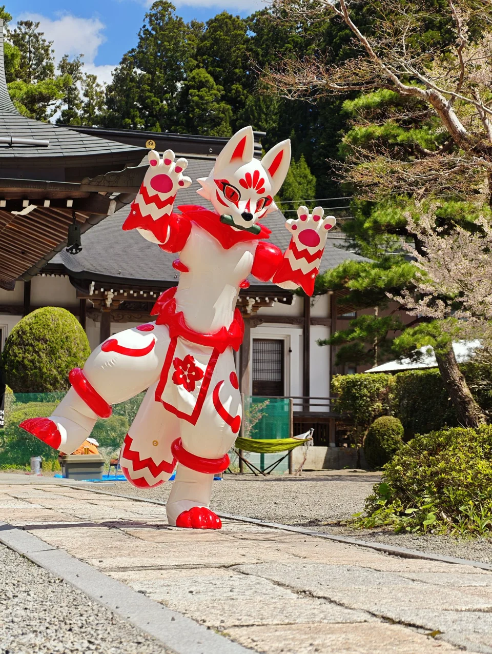 White GUO series latex kitsune with red markings waving in front of a temple