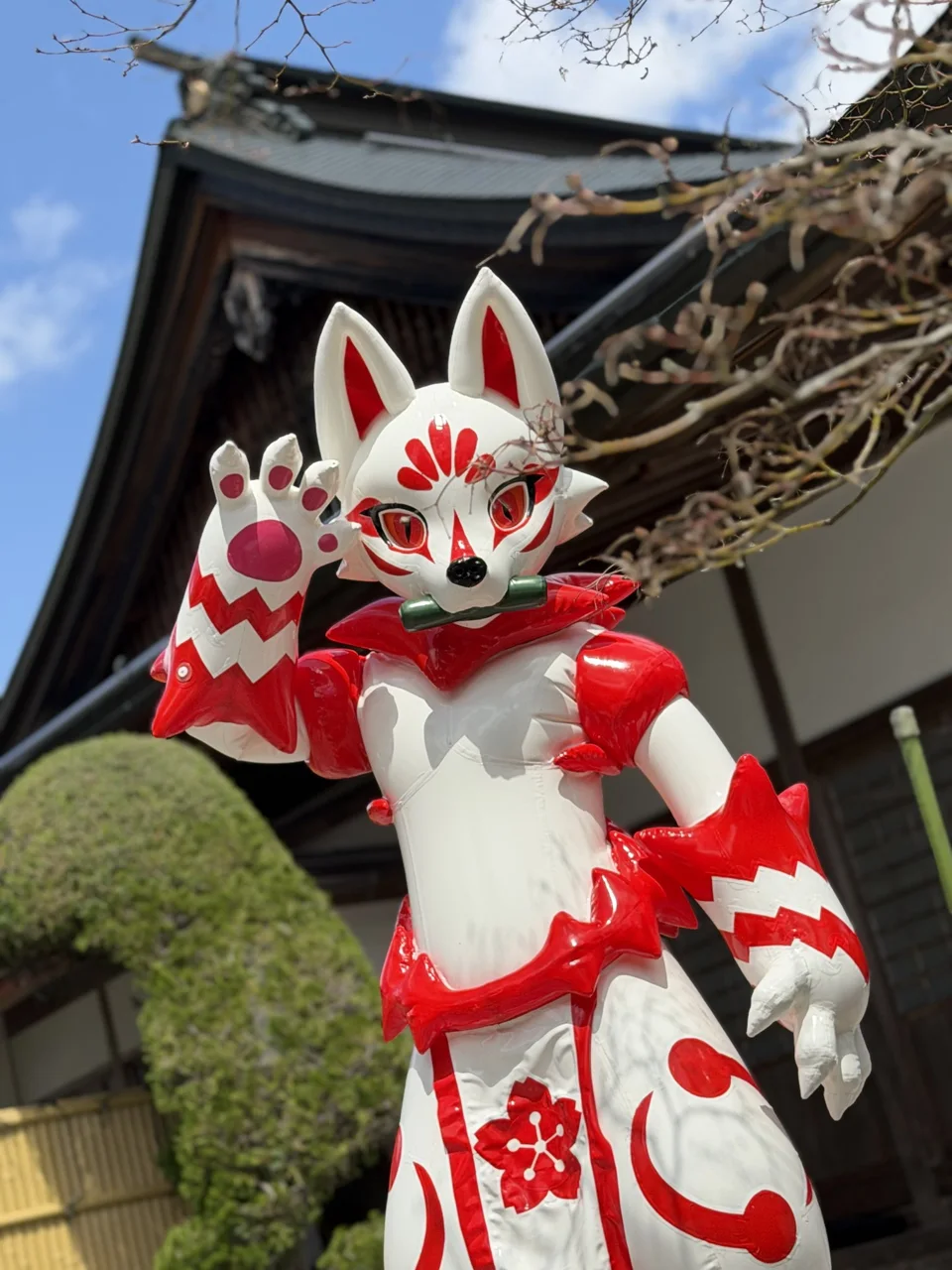 White GUO series latex kitsune with red markings waving in front of a temple